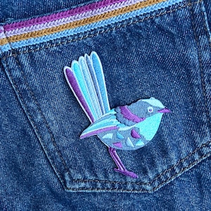May include: A blue and purple embroidered bird patch with a long tail, sewn onto a blue denim pocket.