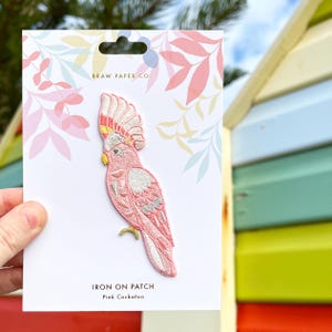 May include: Embroidered pink cockatoo iron-on patch on a white card. The patch shows a detailed design of a pink cockatoo with a yellow crest. The card has the text "BRAW PAPER CO." and "IRON ON PATCH Pink Cockatoo".