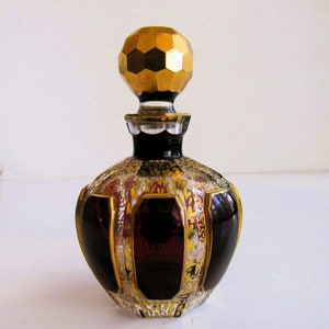 Puede incluir: Una botella de vidrio rojo oscuro con detalles dorados y blancos. La botella tiene un tapón dorado geométrico.