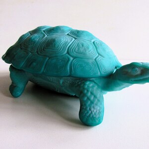 May include: A teal blue glass turtle figurine with a detailed shell and a closed mouth. The turtle is sitting on a white surface.