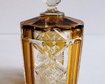 May include: A clear glass jar with a faceted design and amber accents. The jar has a clear glass lid with a faceted knob.