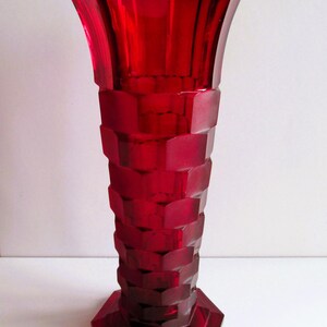 May include: A tall, ruby-red glass vase with a geometric, Art Deco design. The vase features a hexagonal base and a flared top. The body is composed of stacked, rectangular blocks. The vase is approximately 12 inches tall.