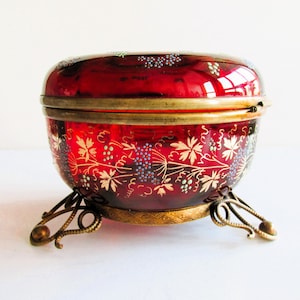 Könnte beinhalten: Eine rote Glas-Schmuckdose mit goldenem Deckel und floralem Design. Die Dose steht auf drei goldenen Metallbeinen.