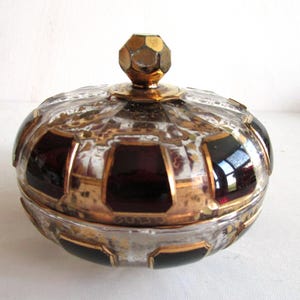 Può includere: Un portagioie vintage con corpo in vetro trasparente, finiture dorate e pannelli rosso scuro. Il coperchio è sormontato da un pomello sfaccettato color oro. Il piatto ha una forma arrotondata ed è probabilmente utilizzato per riporre piccoli oggetti.