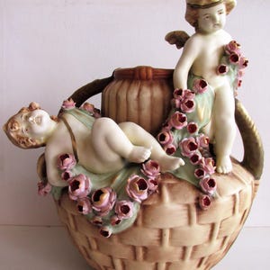 Op de afbeelding: Decoratieve keramische vaas met twee cherubijnachtige figuren en een mandweefpatroon. De figuren zijn versierd met roze rozen en groene bladeren. De vaas heeft een lichtbruine kleur met gouden accenten.