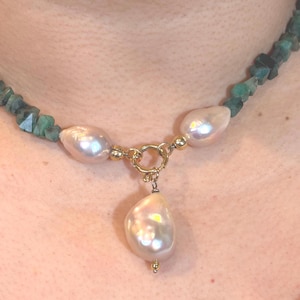 May include: A necklace featuring three large, lustrous pearls suspended from a gold clasp. The necklace is strung with small, faceted, emerald-green gemstones. The pearls have a creamy white color and a unique, irregular shape.