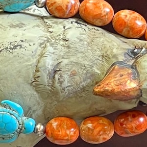May include: A beaded bracelet featuring orange oval-shaped beads and turquoise turtle-shaped beads. The bracelet is displayed on a light green, textured surface. The beads are strung together with silver-colored metal accents.