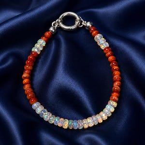 May include: A beaded bracelet featuring alternating sections of orange-red and iridescent opal beads. The bracelet is secured with a silver-colored clasp. The jewelry is displayed on a dark blue satin surface.