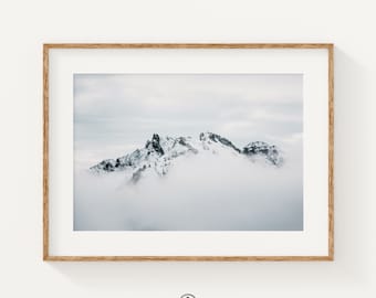 May include: A photograph of snow-capped mountains partially obscured by clouds. The mountains are a dark gray color and the clouds are a light gray color. The sky is a light blue color.