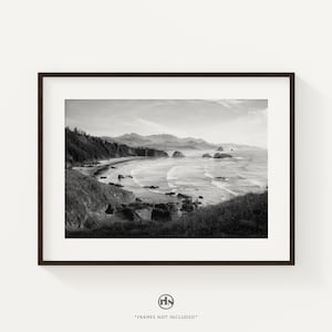 May include: Black and white framed print of a coastal landscape. The image captures a beach with waves, rocks, and a mountainous backdrop. The print is in a dark brown frame and is displayed against a white background.
