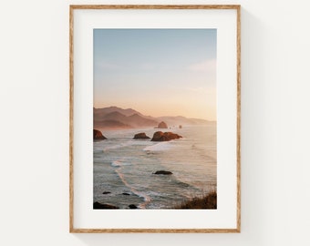 May include: A photograph of a seascape with a mountain range in the background. The ocean is a light blue color with white waves crashing on the shore. The sky is a light blue color with a pink hue at the horizon. The mountains are a dark brown color.