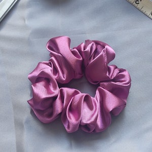 May include: A close-up shot of a scrunchie made of shiny, mauve-colored satin fabric. The scrunchie is circular and gathered, with multiple folds and pleats. The background is a solid, light gray fabric.
