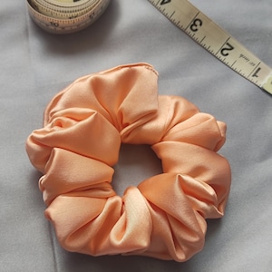 Puede incluir: Una scrunchie de satén color melocotón, un accesorio para el cabello, es el punto focal de la imagen. La scrunchie está hecha de una tela suave y brillante. Una cinta métrica es parcialmente visible en el fondo. La scrunchie es un accesorio elegante para el cabello.