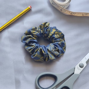 Puede incluir: Una scrunchie azul y dorada estampada sobre una superficie gris, junto a un lápiz amarillo, una cinta métrica y unas tijeras. La scrunchie es de tela y tiene forma circular.