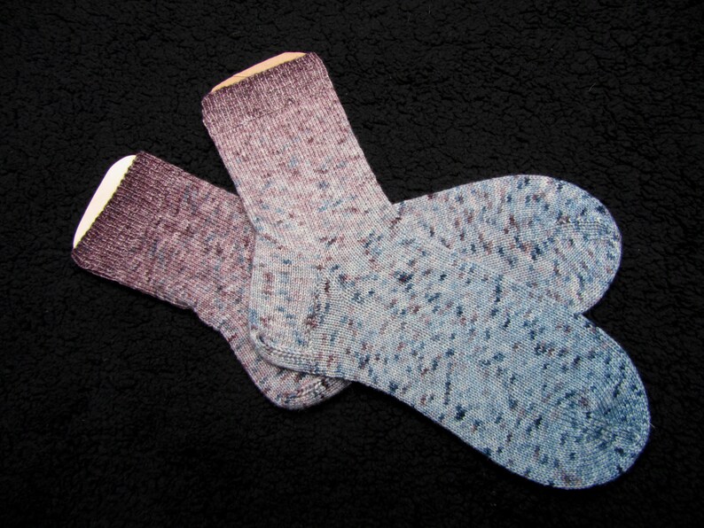 Op de afbeelding: Twee paar sokken in een blauw en paars gespikkeld patroon. De sokken liggen op een zwart, pluche oppervlak.