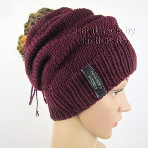Könnte beinhalten: Burgunderfarbene Strickmütze mit Rippenmuster. Die Mütze hat eine lockere Passform und ein kleines schwarzes Etikett mit der Aufschrift "handmade by knitbits.de". Die Mütze wird auf einem Schaufensterkopf gezeigt.