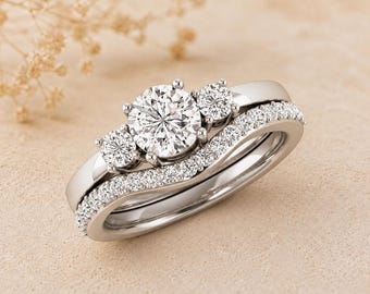 May include: A three-stone engagement ring set with a matching band. The rings are silver-toned, with a large, round center stone and two smaller stones. The band is curved and set with small, sparkling stones.