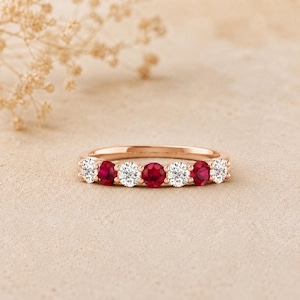 May include: A rose gold band ring featuring an alternating pattern of three round brilliant-cut red rubies and four round brilliant-cut white diamonds set in a shared prong setting, creating a continuous row of seven sparkling gemstones.