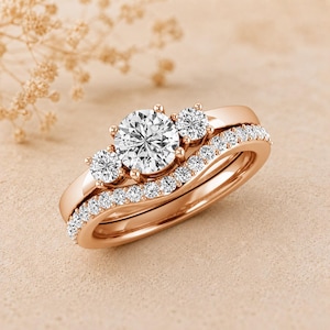 Puede incluir: Juego de anillos de compromiso de oro rosa con un diseño de tres piedras, una piedra central grande y dos piedras laterales más pequeñas. La banda a juego está adornada con una fila de piedras de acento más pequeñas. Los anillos están sobre una superficie neutra.