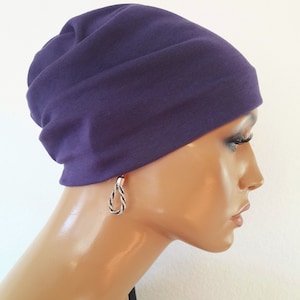 Op de afbeelding: Een dieppaarse beanie-muts van zachte stof. De hoed wordt getoond op een mannequinhoofd, gecombineerd met zilveren en zwarte oorbellen in touwstijl. De achtergrond is effen wit.