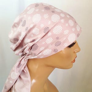 May include: A pink head covering with a pattern of white and gray striped circles. The fabric is gathered at the back and tied, with the ends of the fabric hanging down. The head covering is worn on a mannequin head.