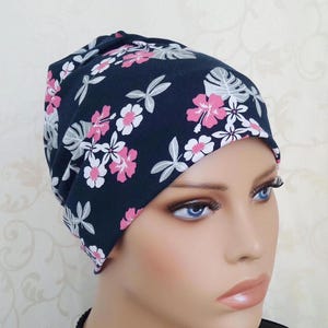 Könnte beinhalten: Eine marineblaue Beanie-Mütze mit einem Blumenmuster aus rosa und weißen Blüten und grauen Blättern. Die Mütze ist aus einem weichen, dehnbaren Stoff gefertigt und passt sich der Kopfform an.