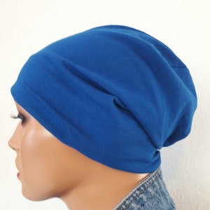 May include: A royal blue beanie hat made of soft fabric. The hat is shown on a mannequin head, with a denim jacket visible at the bottom. The hat is a solid color and has a simple, casual design.