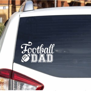 May include: White vinyl decal with the text "Football Dad" and a football graphic. The decal is on the back window of a white car.
