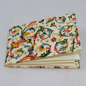 Op de afbeelding: Een decoratief notitieboek met een crèmekleurige omslag met een kleurrijk bloemmotief in rood, geel, blauw en groen. Het notitieboek is open, met lege pagina's. De randen van de pagina's zijn zichtbaar en de omslag is licht gekanteld.