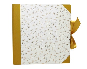 Puede incluir: Álbum de fotos cuadrado con una cubierta blanca con un patrón de semillas de diente de león. El lomo y los detalles de las esquinas son dorados, y una cinta dorada cierra el álbum. Diseñado para guardar fotografías.