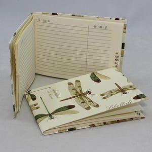 May include: An open address book with lined pages and a dragonfly-patterned cover. The cover features illustrations of dragonflies in shades of brown, green, and beige. The book is open to reveal lined pages for names and addresses.