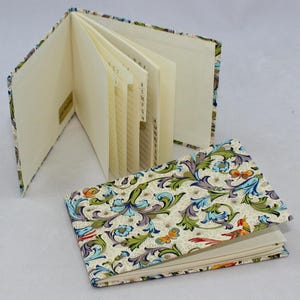 May include: Two address books, one open and one closed, featuring a floral pattern in green, blue, and purple on a cream background. The open book has tabbed pages. The closed book displays the cover design with butterflies and birds.