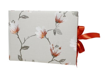 May include: A white fabric photo album with a floral pattern in shades of orange, gray, and white. The album has a red ribbon tied around it.