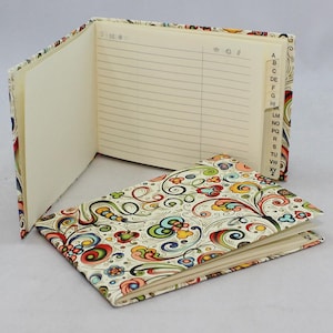 May include: An open address book with lined pages and an alphabetized tab index. The cover and spine feature a colorful floral pattern with swirls and various shapes in shades of red, yellow, green, and blue on a cream background.