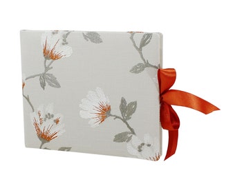 May include: A rectangular photo album with a floral pattern. The cover is a light beige with white and orange flowers and gray leaves. An orange ribbon is tied on the side. The album is closed.