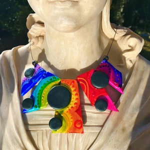 Może przedstawiać: Kolorowy, abstrakcyjny naszyjnik o geometrycznych kształtach i tęczowej kolorystyce. Naszyjnik posiada czarne okrągłe akcenty i jest nawleczony na cienki czarny sznurek. Projekt zawiera odcienie czerwieni, pomarańczu, żółci, zieleni, niebieskiego i fioletu.