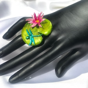 Può includere: Un anello verde e dorato con un giglio d'acqua rosa e una libellula blu in cima.