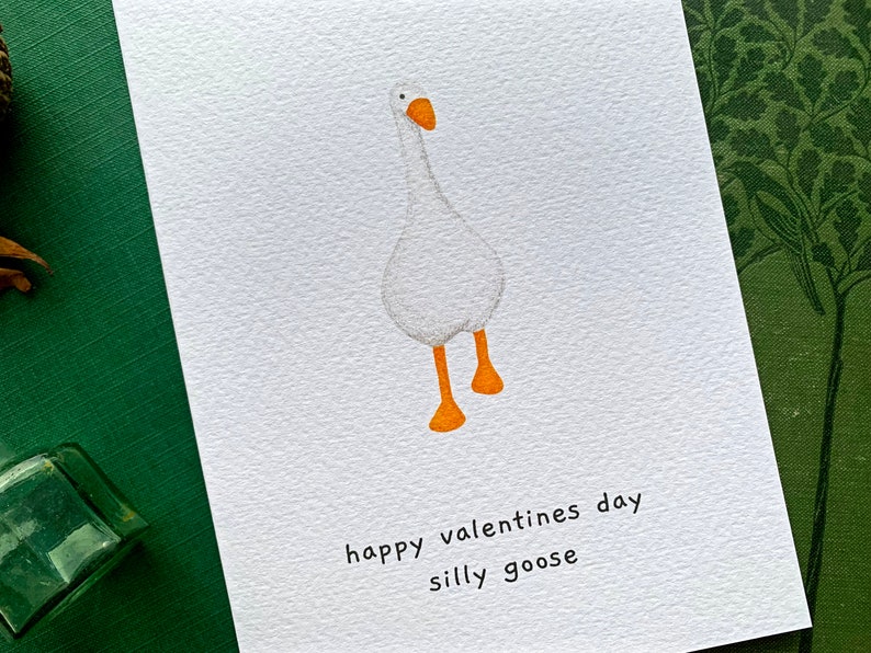 May include: A white card featuring a cartoon goose with orange feet and beak. The text "happy valentines day silly goose" is printed below the illustration. The card is set against a green backdrop.