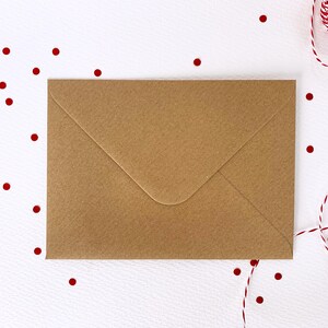 May include: A brown paper envelope lies flat on a white surface, with red confetti dots scattered around it. A red and white twine is in the upper right corner. The envelope is sealed and ready for mailing.
