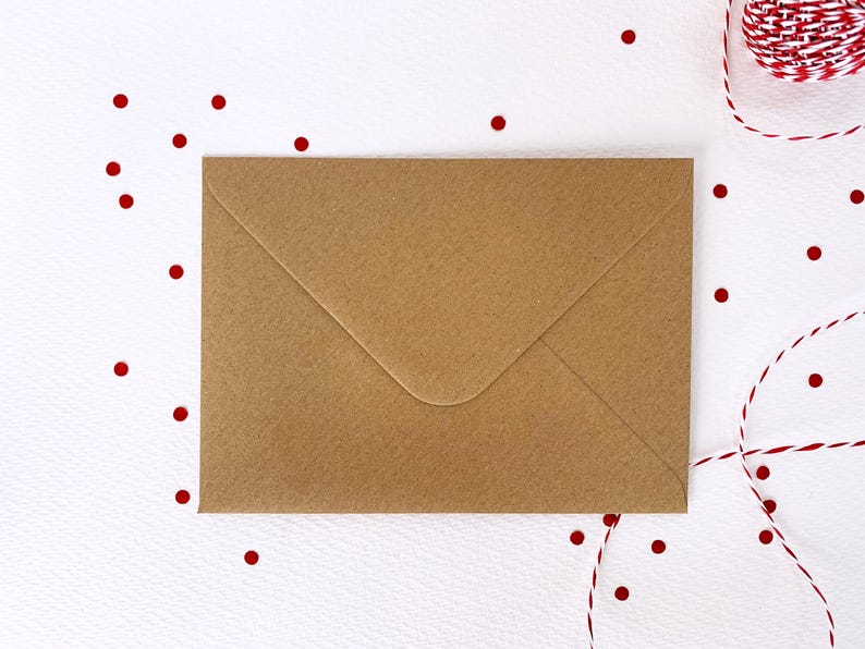 May include: A brown paper envelope lies flat on a white surface, with red confetti dots scattered around it. A red and white twine is in the upper right corner. The envelope is sealed and ready for mailing.