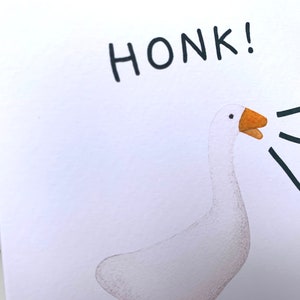 Może przedstawiać: Ilustracja białej gęsi z pomarańczowym dziobem i napisem "HONK!" czarnym tekstem. Czarne linie wychodzą z dzioba gęsi, sugerując dźwięk gęgania. Grafika znajduje się na białym tle.