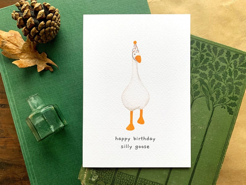 May include: A white goose wearing a party hat with orange and white polka dots. The goose is standing on two orange legs. The text "happy birthday silly goose" is written below the goose.