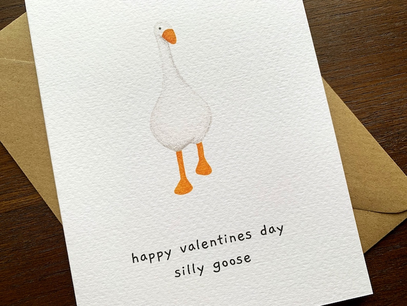May include: A white card featuring a cartoon goose with orange feet and beak. The text "happy valentines day silly goose" is printed below the illustration. The card is accompanied by a brown envelope.