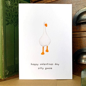 May include: A Valentine's Day card with a watercolour illustration of a white goose with orange feet and beak. The card has the text "happy valentines day silly goose" in black on a white background.