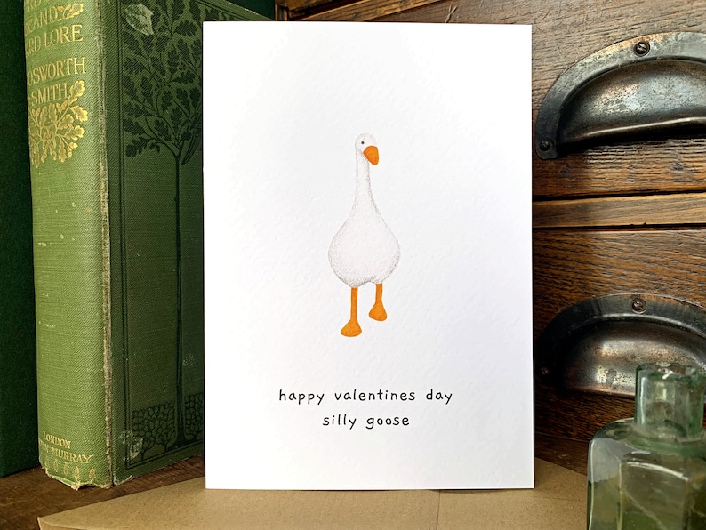 May include: A Valentine's Day card with a watercolour illustration of a white goose with orange feet and beak. The card has the text "happy valentines day silly goose" in black on a white background.