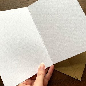May include: A blank greeting card with a textured white interior, held by a hand. A brown envelope is visible in the background, suggesting a stationery item for sending messages or notes.