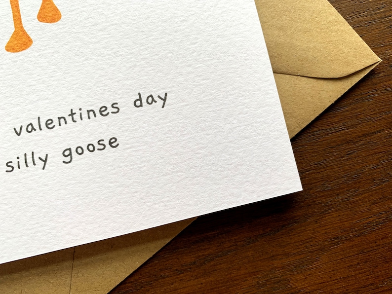May include: A white greeting card with the text "valentines day silly goose" in black. The card is paired with a brown envelope and rests on a dark wood surface. Orange decorative elements are visible at the top.