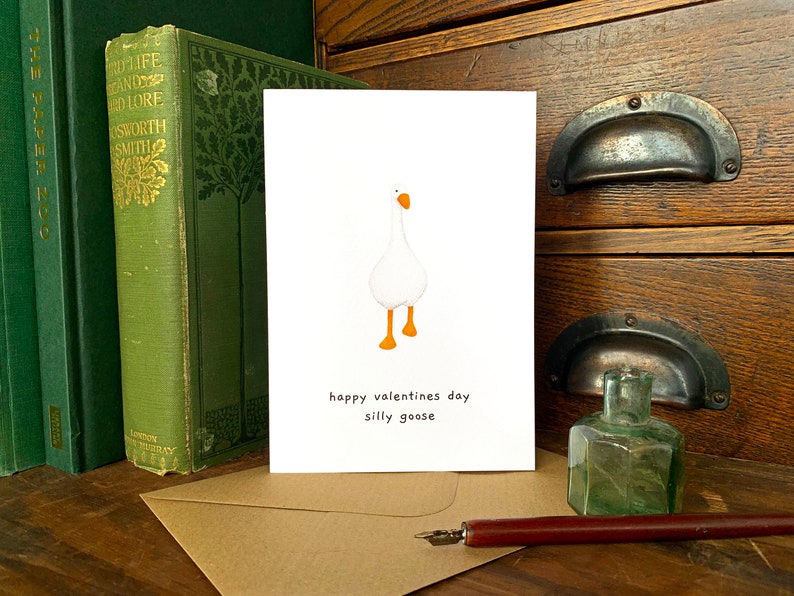 May include: A white card featuring a cartoon goose with orange feet and beak. The text on the card reads "happy valentines day silly goose". The card is on a wooden surface, with green books and an inkwell visible in the background.