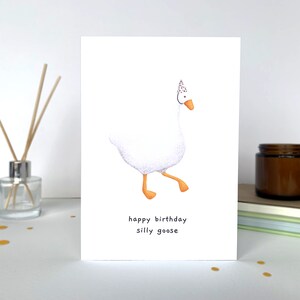 K&ouml;nnte beinhalten: Eine wei&szlig;e Gans mit einer roten und wei&szlig;en Partym&uuml;tze mit Punkten geht nach rechts. Der Text "happy birthday silly goose" befindet sich unter der Gans.