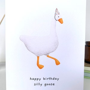 K&ouml;nnte beinhalten: Eine wei&szlig;e Gans mit einem bunten Partyhut und dem Text "happy birthday silly goose" auf wei&szlig;em Hintergrund.